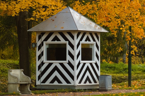 A small, striped black and white shelter with a pyramidal roof is set amidst a backdrop of vibrant yellow autumn leaves. There is a stone bench to the left and a small metal bin nearby, both resting on a paved surface. The lush greenery and trees surround the structure, contributing to an overall pastoral setting.