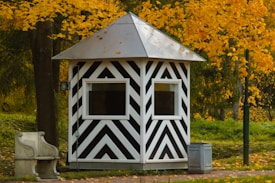 A small, striped black and white shelter with a pyramidal roof is set amidst a backdrop of vibrant yellow autumn leaves. There is a stone bench to the left and a small metal bin nearby, both resting on a paved surface. The lush greenery and trees surround the structure, contributing to an overall pastoral setting.