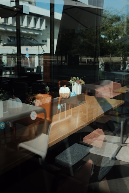 A wooden table positioned in a modern coffee shop is adorned with decorative elements, including small potted plants and a stylized white pumpkin. The interior is lit by natural light filtering through large windows, with reflections of the exterior buildings visible on the glass. Various chairs are arranged around the table, creating a cozy and inviting atmosphere.