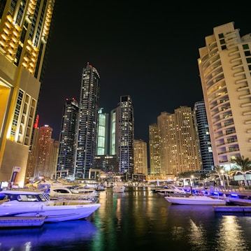 A vibrant cityscape at night with towering skyscrapers illuminated by lights. The foreground features a marina with several yachts docked in the water, reflecting the glow of the city lights. The scene conveys a sense of luxury and modernity, with the tall buildings creating an impressive backdrop.