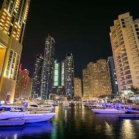 A vibrant cityscape at night with towering skyscrapers illuminated by lights. The foreground features a marina with several yachts docked in the water, reflecting the glow of the city lights. The scene conveys a sense of luxury and modernity, with the tall buildings creating an impressive backdrop.