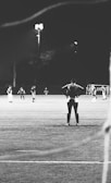 A black and white photograph of a nighttime soccer match, showing several players on the field. One player stands in the foreground with hands on hips, suggesting a moment of reflection or anticipation. A lit field in the background provides contrast to the dark surroundings.
