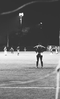 A black and white photograph of a nighttime soccer match, showing several players on the field. One player stands in the foreground with hands on hips, suggesting a moment of reflection or anticipation. A lit field in the background provides contrast to the dark surroundings.