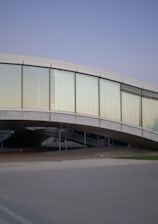 A modern architectural structure featuring a curved glass facade with large windows set within a gentle arc. The building's design incorporates smooth concrete elements and an elevated pathway underneath, creating a sense of openness. The sky is a clear, pale blue, contributing to a serene environment.