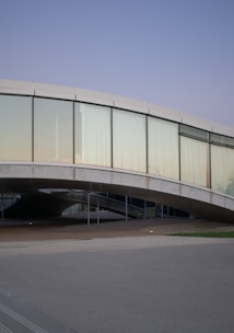 A modern architectural structure featuring a curved glass facade with large windows set within a gentle arc. The building's design incorporates smooth concrete elements and an elevated pathway underneath, creating a sense of openness. The sky is a clear, pale blue, contributing to a serene environment.