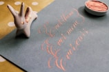 A calligraphy artwork on a blue sheet of paper, showing the words 'Ever thine, Ever mine, Ever yours', written in elegant pink ink. To the left, there is a small ceramic rabbit figurine, and at the top right, an open container with pink ink or paint.