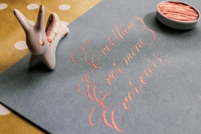 A calligraphy artwork on a blue sheet of paper, showing the words 'Ever thine, Ever mine, Ever yours', written in elegant pink ink. To the left, there is a small ceramic rabbit figurine, and at the top right, an open container with pink ink or paint.