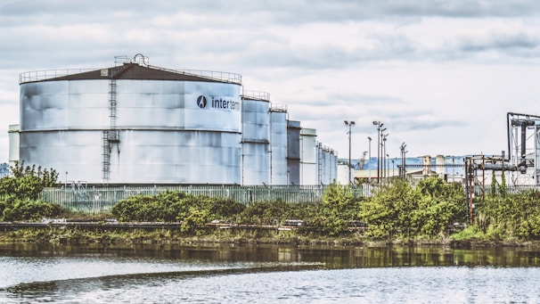 Large industrial storage tanks are situated near a water body, surrounded by greenery and a fence. The tanks are metallic and cylindrical with visible infrastructure and pipes. The sky is overcast with clouds, adding a muted tone to the scene.