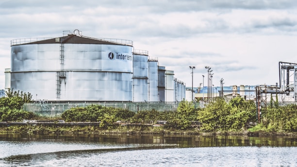 Large industrial storage tanks are situated near a water body, surrounded by greenery and a fence. The tanks are metallic and cylindrical with visible infrastructure and pipes. The sky is overcast with clouds, adding a muted tone to the scene.