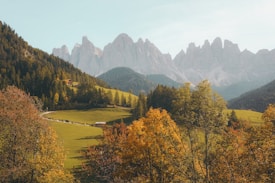 A picturesque landscape features rolling green hills dotted with trees displaying vibrant autumn colors. In the background, majestic mountains rise under a clear blue sky, and a few small wooden cabins are nestled among the trees.