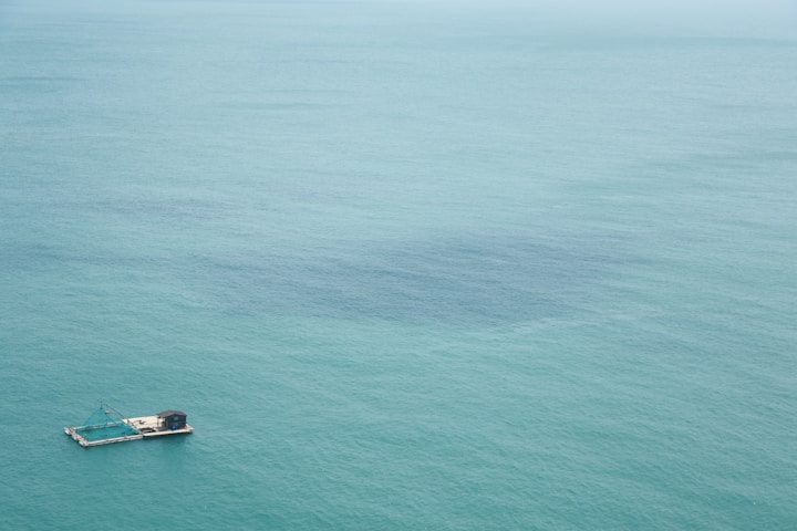 A lone floating platform with a structure on it is situated in the open sea. The water is a calm, expansive teal blue color, stretching out to the horizon.