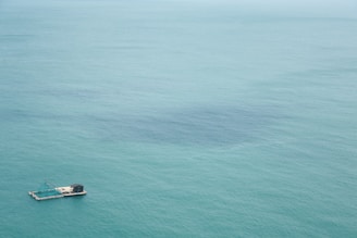 A lone floating platform with a structure on it is situated in the open sea. The water is a calm, expansive teal blue color, stretching out to the horizon.