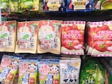 Various packaged snacks and candies are displayed on a store shelf. The packages feature colorful designs with images of fruits, beverages, and cartoon characters. The text on the packaging is in Japanese, with prominent depictions of strawberries, matcha, and milk tea.
