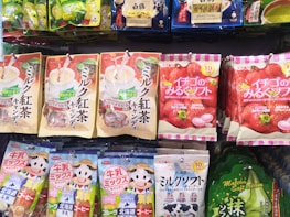 Various packaged snacks and candies are displayed on a store shelf. The packages feature colorful designs with images of fruits, beverages, and cartoon characters. The text on the packaging is in Japanese, with prominent depictions of strawberries, matcha, and milk tea.