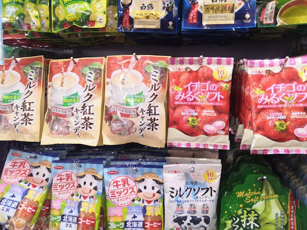 Various packaged snacks and candies are displayed on a store shelf. The packages feature colorful designs with images of fruits, beverages, and cartoon characters. The text on the packaging is in Japanese, with prominent depictions of strawberries, matcha, and milk tea.