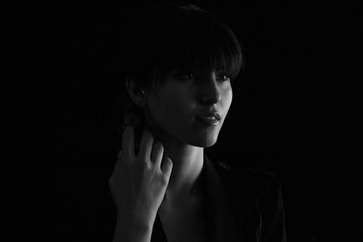 A woman is portrayed in a dramatic black and white setting with soft lighting casting delicate shadows across her face. She has short hair and is touching her neck with one hand. The dark background adds to the intensity of the image.