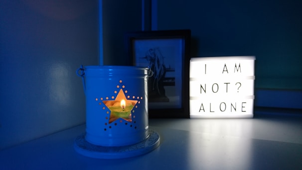 A dimly lit scene featuring a white lantern with a star-shaped cutout, casting a warm glow from a candle inside. Next to it, a lightbox displays the words 'I AM NOT? ALONE' in black letters. A framed art piece is partially visible behind the lightbox. The environment is infused with a blue tone, creating a calm and introspective atmosphere.