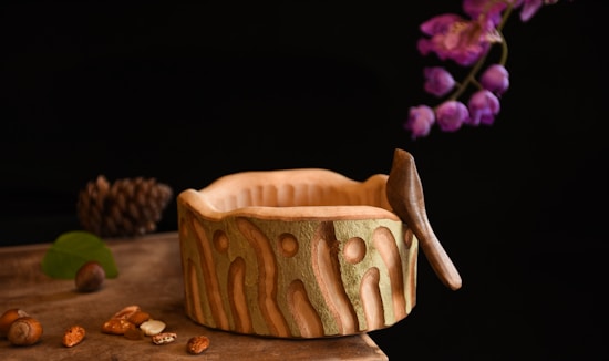 A wooden bowl with intricate carvings sits on a wooden surface alongside scattered nuts and green leaves. A small carved bird perches on the edge of the bowl. In the background, purple flowers drape elegantly, adding a touch of color to the overall warm and earthy composition.