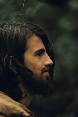 A close-up profile of a man with long, dark hair and a beard, set against a blurred natural background. The lighting gives the image a warm, earthy tone, highlighting features and creating a serene atmosphere.