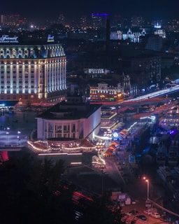 A nighttime cityscape featuring a brightly lit hotel with the name Fairmont Grand Hotel visible. The street below is decorated with lights and lined with colorful stalls, possibly part of a market or festival. Light trails from vehicles can be seen on the road, and the skyline is dotted with buildings illuminated against the night sky.