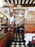 An interior of a vintage-style restaurant with a classic decor, featuring ornate wooden furnishings, a checkered floor, and a staircase adorned with traditional ceramic tiles. Various decorative plates, paintings, and certificates are displayed on the walls. A person is seen ascending the staircase.