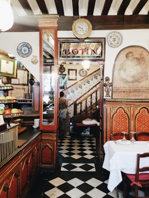 An interior of a vintage-style restaurant with a classic decor, featuring ornate wooden furnishings, a checkered floor, and a staircase adorned with traditional ceramic tiles. Various decorative plates, paintings, and certificates are displayed on the walls. A person is seen ascending the staircase.