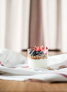 A glass filled with layers of creamy yogurt, granola, fresh blueberries, and sliced strawberries on top. The dessert is placed on a white cloth with red stripes, against a soft-focused background.
