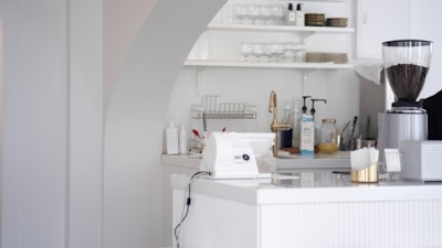 A white, modern kitchen counter area with various objects. There is a white card payment machine on the counter. Shelves behind the counter hold glasses and plates. An electric grinder containing coffee beans is seen on the right side, and various kitchen supplies, including a bottle of hand sanitizer, are scattered on the surface.