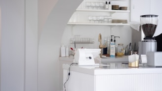 A white, modern kitchen counter area with various objects. There is a white card payment machine on the counter. Shelves behind the counter hold glasses and plates. An electric grinder containing coffee beans is seen on the right side, and various kitchen supplies, including a bottle of hand sanitizer, are scattered on the surface.