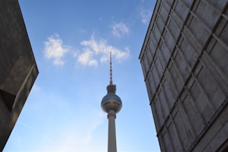 A tall telecommunications tower stands prominently against a clear blue sky, flanked by two modern buildings with a mix of concrete and glass surfaces. The perspective creates a sense of looking up towards the sky.