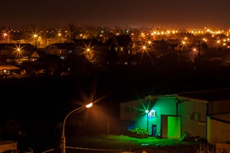 A nighttime view of a residential area with numerous streetlights emitting a warm orange glow, creating a starburst effect in the darkness. The foreground features a building illuminated by a striking green light, contrasting with the otherwise dark environment. In the distance, the silhouettes of houses and trees are visible beneath a cloudy sky.