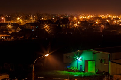 A nighttime view of a residential area with numerous streetlights emitting a warm orange glow, creating a starburst effect in the darkness. The foreground features a building illuminated by a striking green light, contrasting with the otherwise dark environment. In the distance, the silhouettes of houses and trees are visible beneath a cloudy sky.