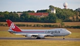 A large cargo airplane on a runway with a landscape of fields, trees, and buildings in the background. The plane has a red tail with a logo and the word 'cargolux' on its body.