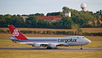 A large cargo airplane on a runway with a landscape of fields, trees, and buildings in the background. The plane has a red tail with a logo and the word 'cargolux' on its body.