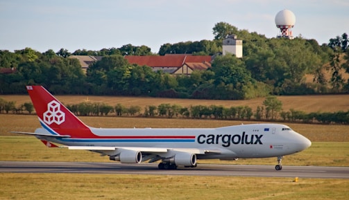 A large cargo airplane on a runway with a landscape of fields, trees, and buildings in the background. The plane has a red tail with a logo and the word 'cargolux' on its body.