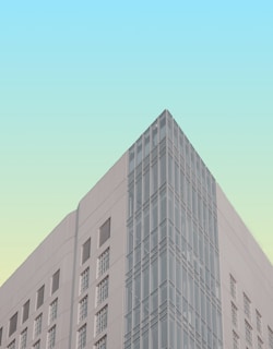 A modern building with a geometric design featuring a combination of large windows and solid walls. The facade includes a corner section with floor-to-ceiling glass panels, allowing for a reflective quality. The background sky has a gradient from light blue to pale green, adding an artistic touch to the scene.