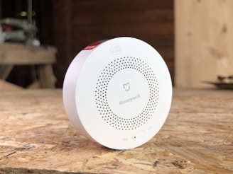 A circular white Honeywell device placed on a wooden surface, possibly a smoke detector or air quality monitor. The device has a series of small holes arranged in a circular pattern and features branding and a small switch on the front. The background is blurred, suggesting an indoor setting with wooden textures.
