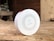 A circular white Honeywell device placed on a wooden surface, possibly a smoke detector or air quality monitor. The device has a series of small holes arranged in a circular pattern and features branding and a small switch on the front. The background is blurred, suggesting an indoor setting with wooden textures.