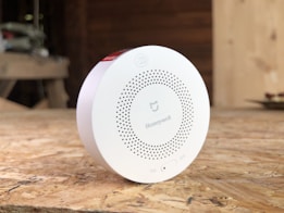 A circular white Honeywell device placed on a wooden surface, possibly a smoke detector or air quality monitor. The device has a series of small holes arranged in a circular pattern and features branding and a small switch on the front. The background is blurred, suggesting an indoor setting with wooden textures.