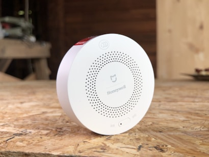A circular white Honeywell device placed on a wooden surface, possibly a smoke detector or air quality monitor. The device has a series of small holes arranged in a circular pattern and features branding and a small switch on the front. The background is blurred, suggesting an indoor setting with wooden textures.