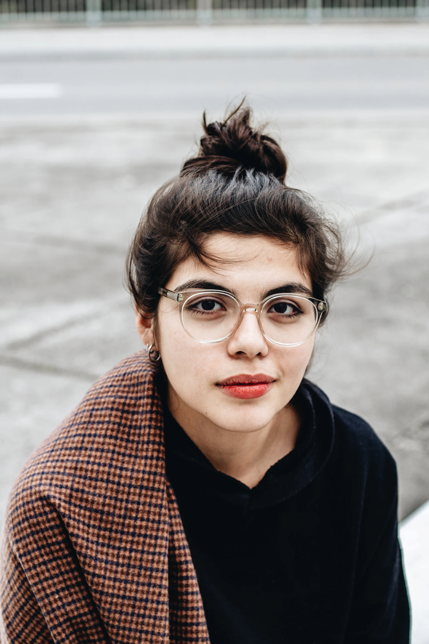 A woman with her hair in a top knot wearing fine round frames on a grey street