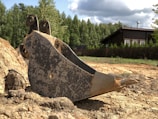 A large metal excavator bucket rests on a dirt ground near a pile of earth. In the background, there is a wooden house with a dark roof surrounded by a tall wooden fence and dense, green trees. The sky is partly cloudy, casting shadows and patches of sunlight on the scene.