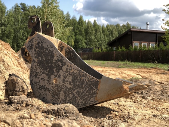 A large metal excavator bucket rests on a dirt ground near a pile of earth. In the background, there is a wooden house with a dark roof surrounded by a tall wooden fence and dense, green trees. The sky is partly cloudy, casting shadows and patches of sunlight on the scene.