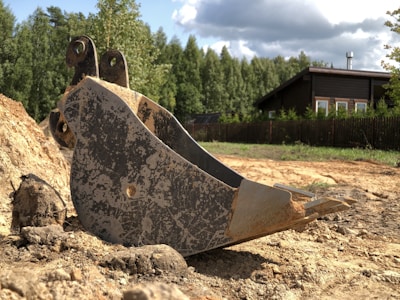 A large metal excavator bucket rests on a dirt ground near a pile of earth. In the background, there is a wooden house with a dark roof surrounded by a tall wooden fence and dense, green trees. The sky is partly cloudy, casting shadows and patches of sunlight on the scene.