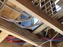 A construction setting with exposed wooden beams and several metal conduits and cables running through them. The conduits are metallic and flexible, suggesting they might be used for electrical wiring or plumbing. Multiple blue and red pipes are visible, likely indicating different utilities. The wood is untreated, indicating an unfinished construction phase.