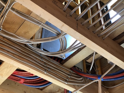 A construction setting with exposed wooden beams and several metal conduits and cables running through them. The conduits are metallic and flexible, suggesting they might be used for electrical wiring or plumbing. Multiple blue and red pipes are visible, likely indicating different utilities. The wood is untreated, indicating an unfinished construction phase.