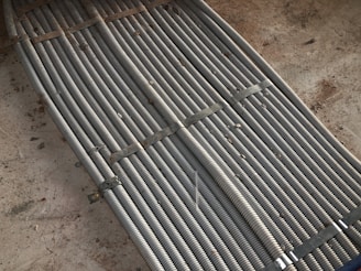 Metal coils are tightly bundled together, secured with brackets, forming a uniform and structured pattern. There is a background of rough concrete with scattered dirt and debris.