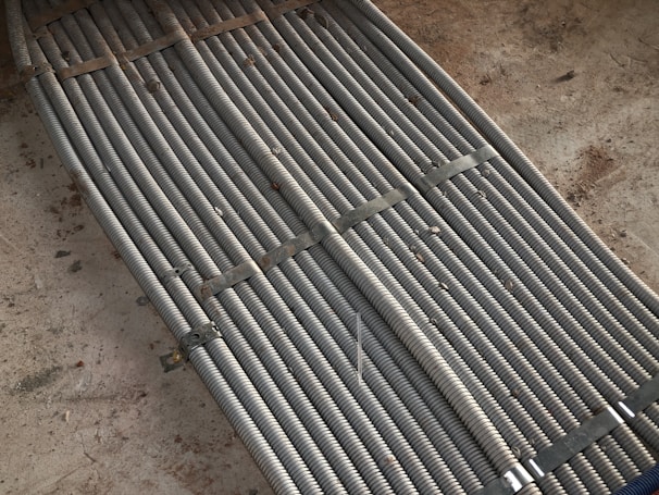 Metal coils are tightly bundled together, secured with brackets, forming a uniform and structured pattern. There is a background of rough concrete with scattered dirt and debris.