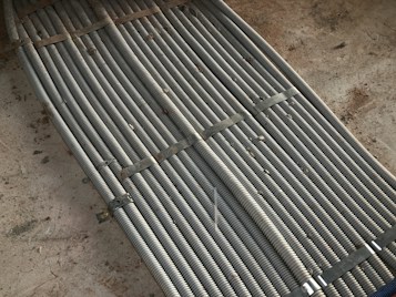 Metal coils are tightly bundled together, secured with brackets, forming a uniform and structured pattern. There is a background of rough concrete with scattered dirt and debris.