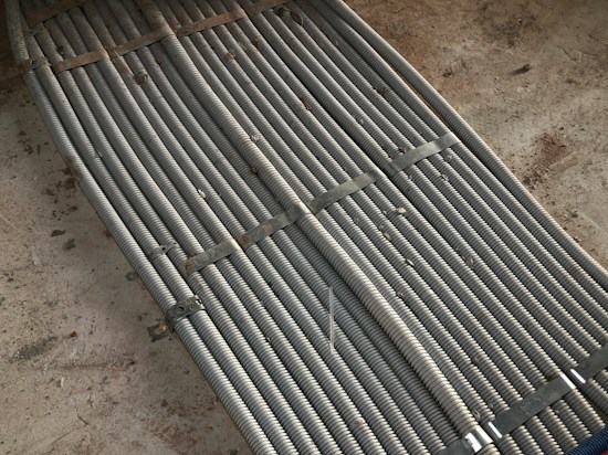 Metal coils are tightly bundled together, secured with brackets, forming a uniform and structured pattern. There is a background of rough concrete with scattered dirt and debris.